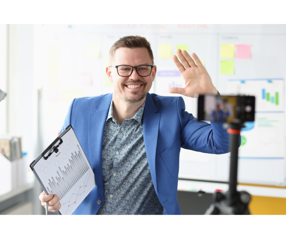 A man sitting down, wearing a blue jacket and glasses, waving to a smartphone on a tripod in front of him